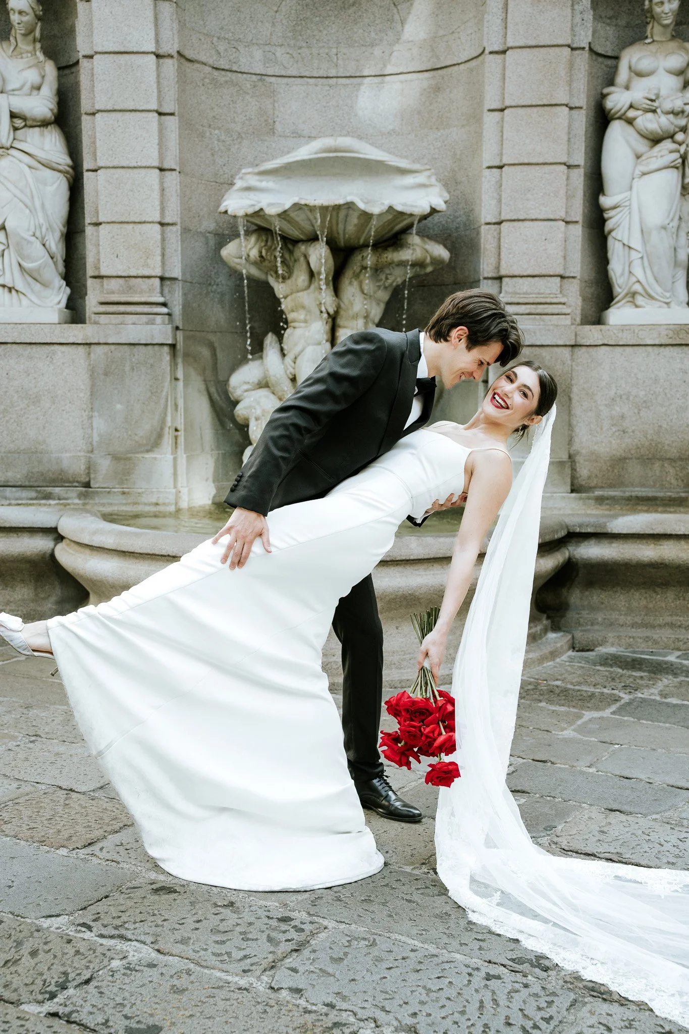 A bride and groom are posing in front of a stone fountain with classical sculptures. The groom is lifting the bride, who is holding a bouquet of red roses, and they are smiling at each other.
