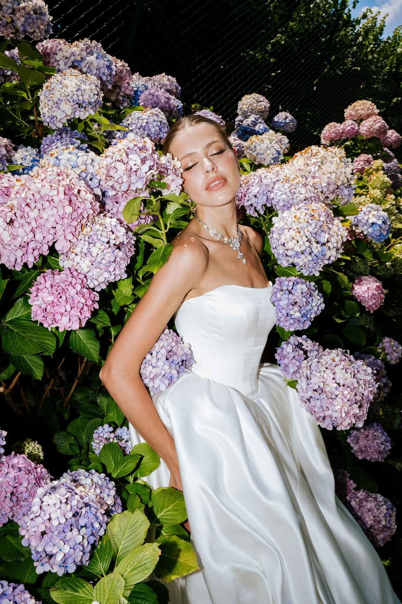 A woman in a white strapless dress posing among pink and purple hydrangea flowers with her eyes closed and head tilted slightly upward.