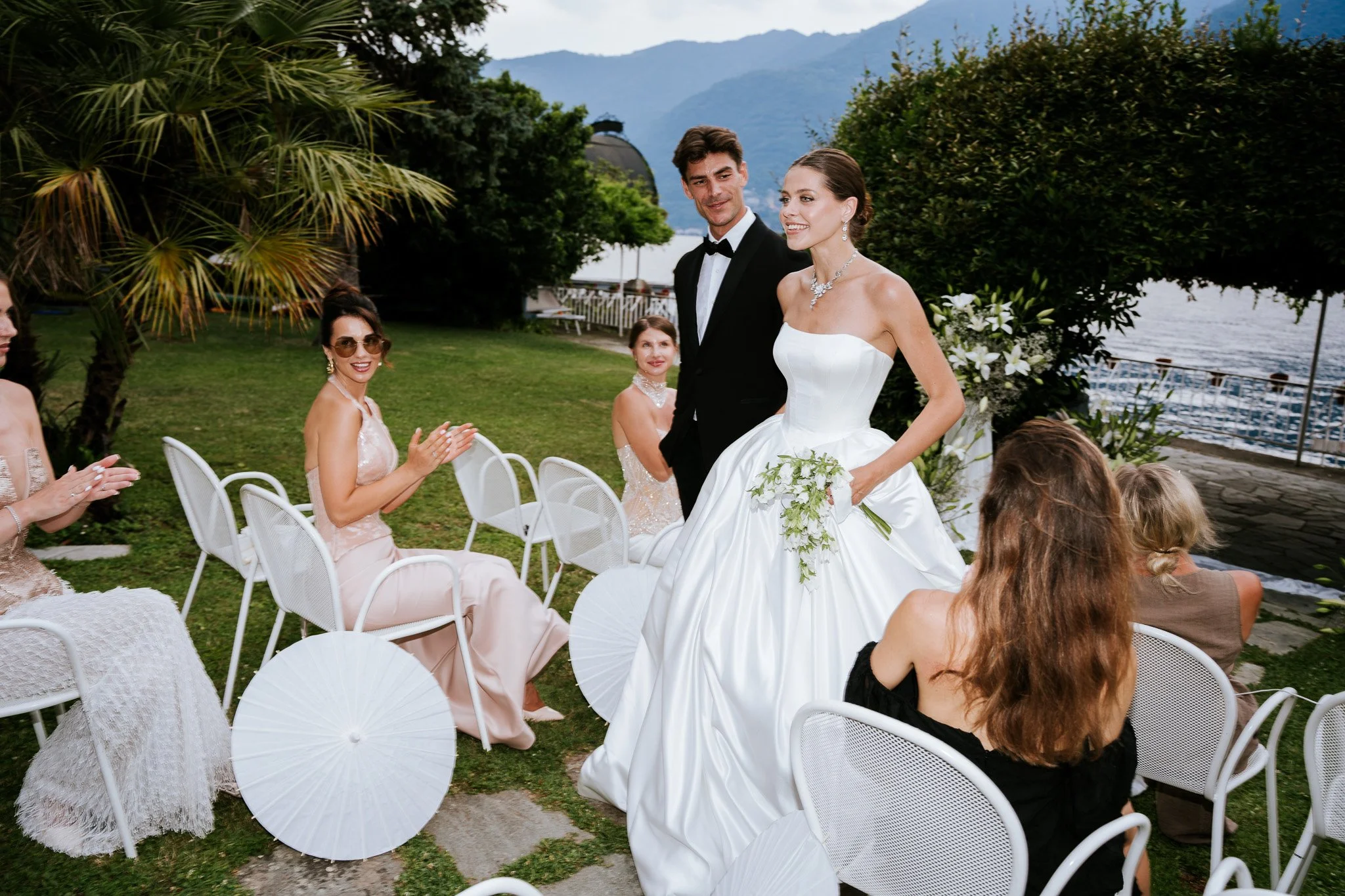 A bride and groom celebrating at a wedding outdoors near a lake with mountains in the background. The bride is smiling, wearing a strapless white wedding gown and jewelry, holding a bouquet. The groom is standing beside her in a black tuxedo. Guests 