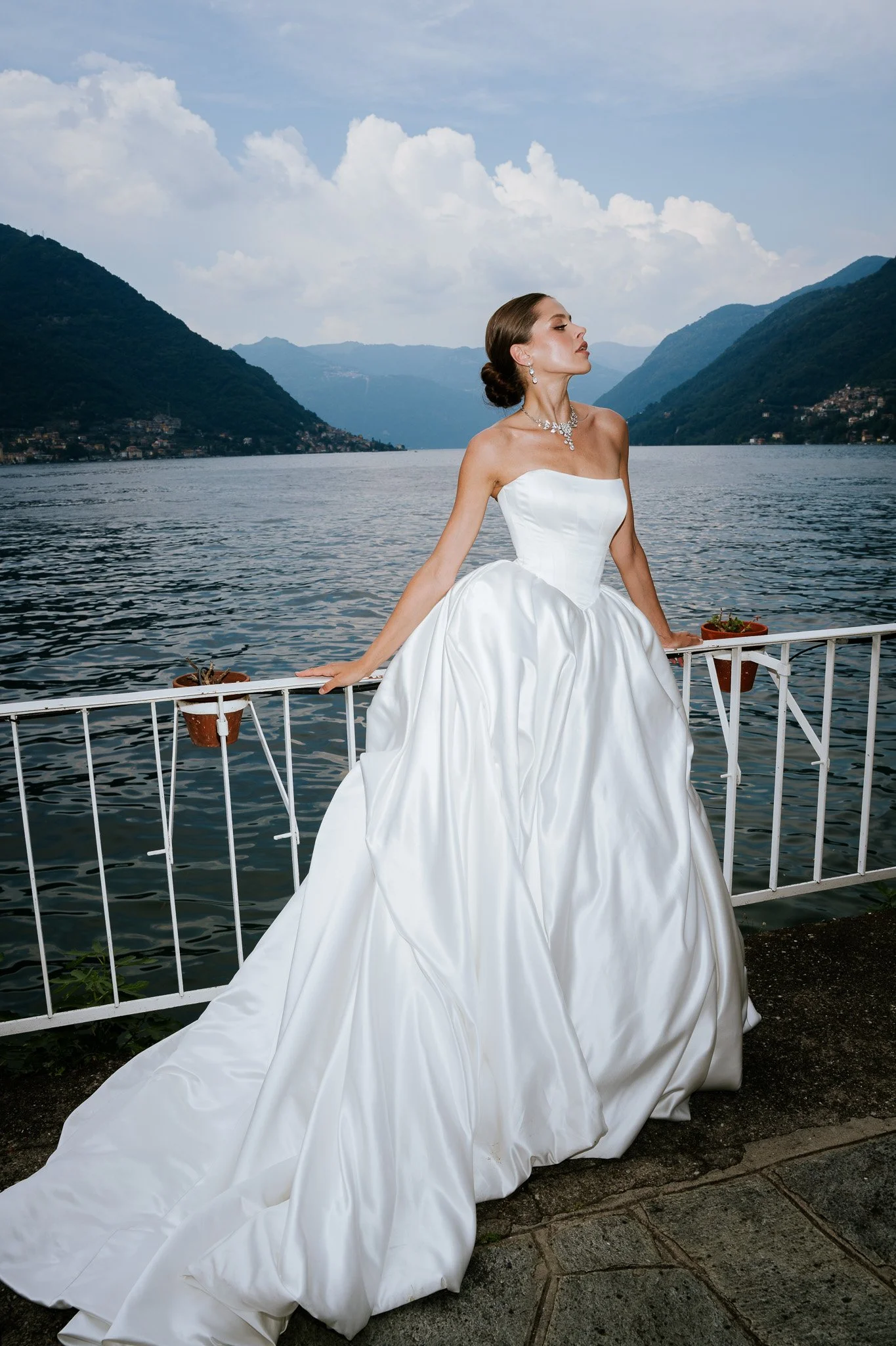 A woman in a white wedding gown and jewelry sits on a white railing near a lake, with mountains and a cloudy sky in the background.