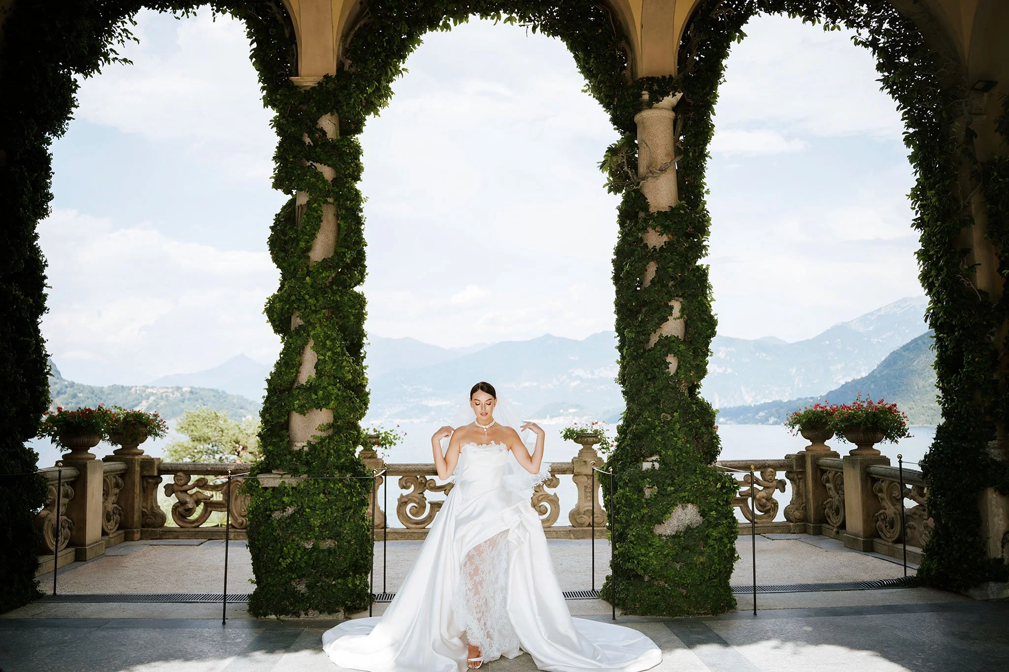 Bride sitting on a stone bench under the ivy-covered loggia arches at Villa del Balbianello, Lake Como, with mountain views behind her.