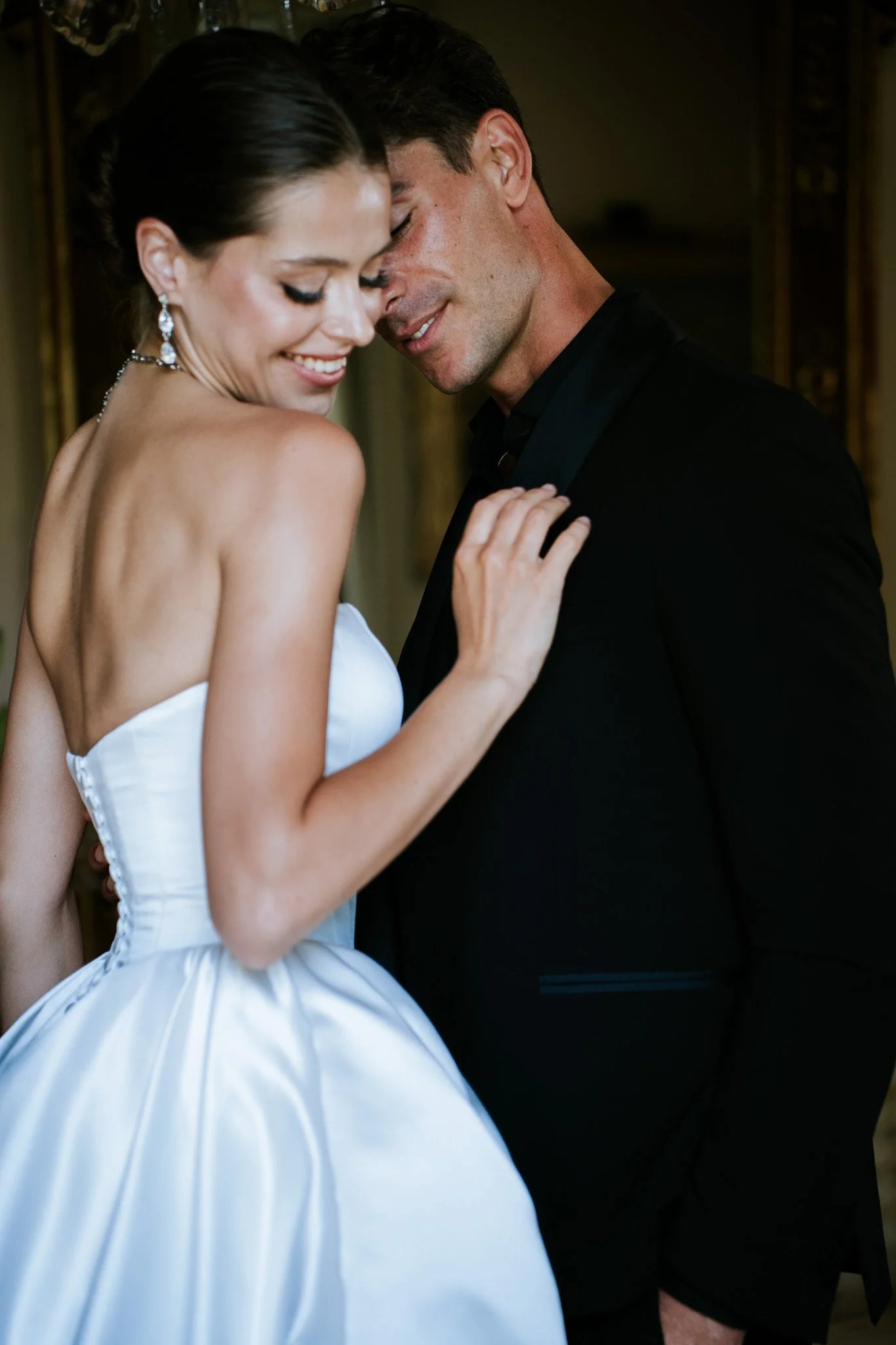 A woman in a white strapless dress, wearing earrings and a necklace, smiling with eyes closed, leaning into a man in a black suit, both with their foreheads and noses touching in an intimate moment.