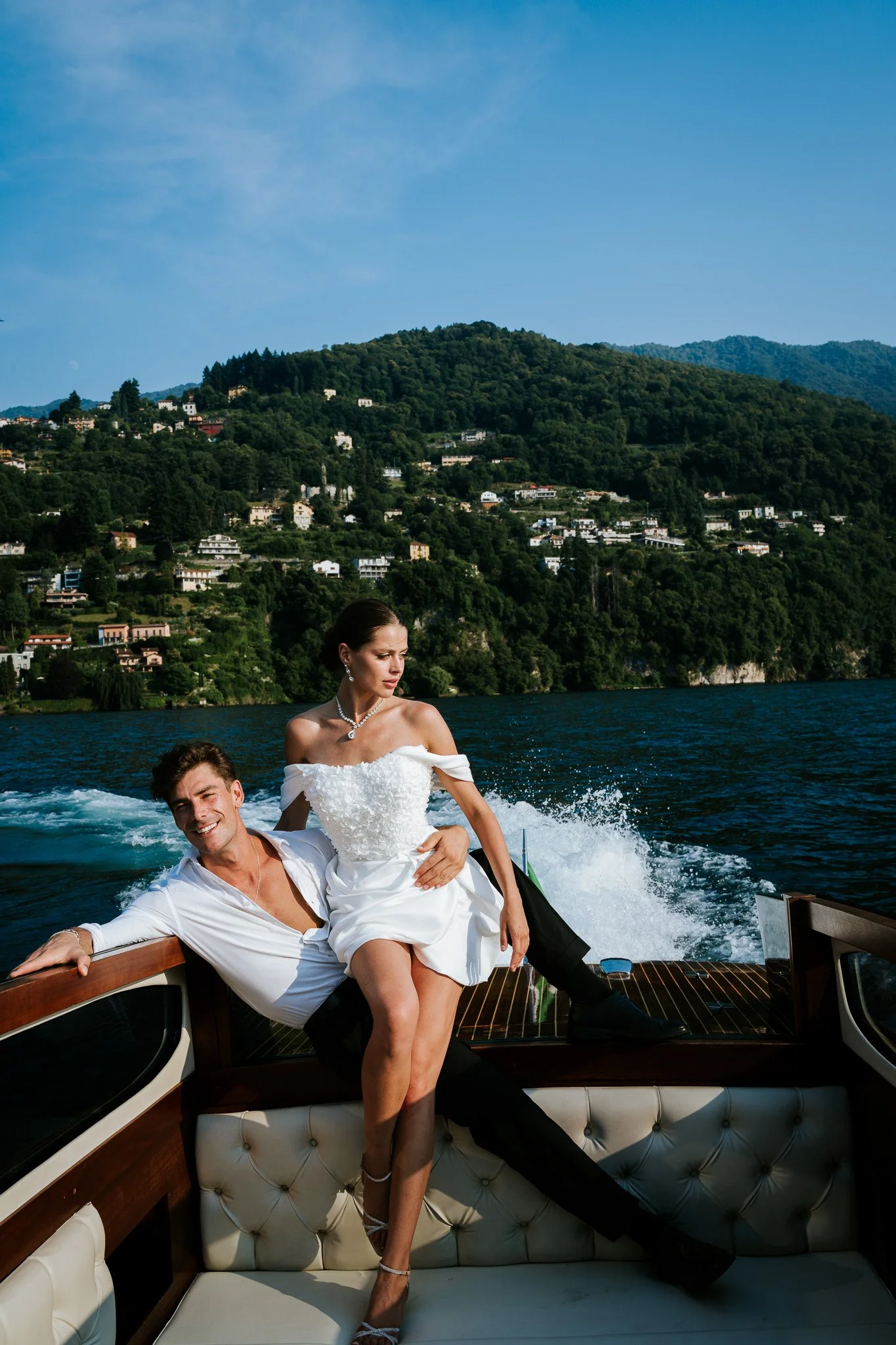 A couple dressed in wedding attire enjoying a boat ride on a lake with green hills and houses in the background.
