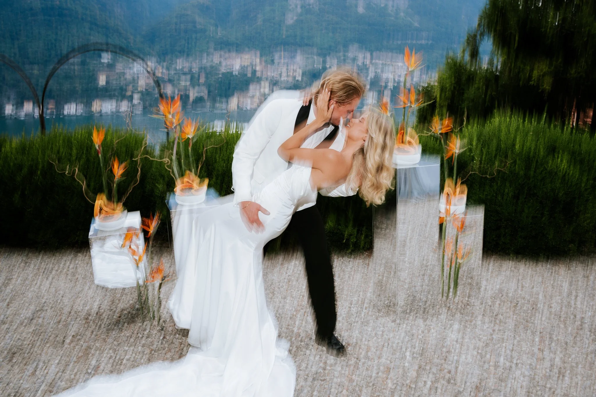 A couple dancing closely, with the woman in a white dress and the man in a white suit, surrounded by orange flowers and modern glass and natural scenery background.