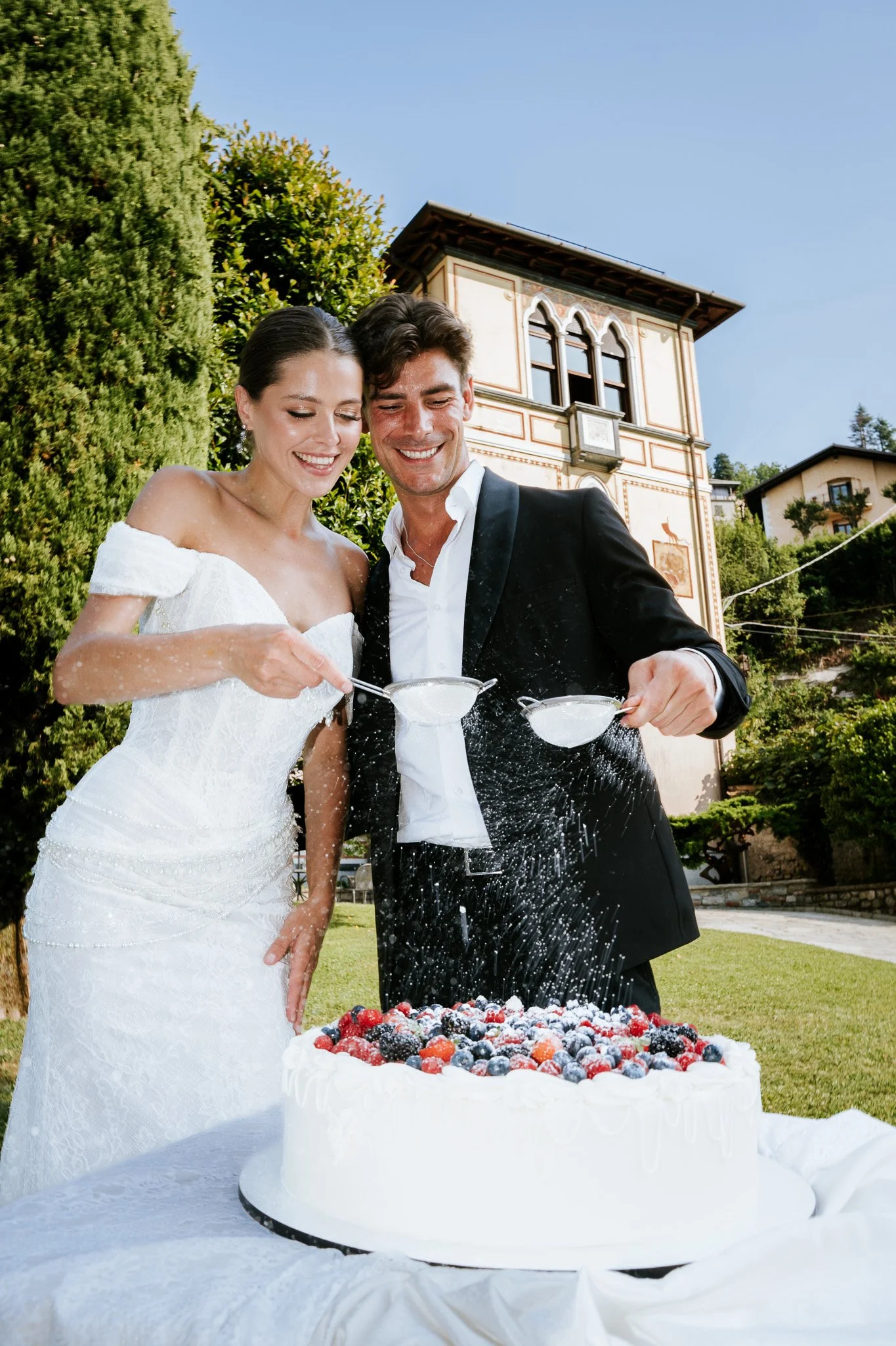 A bride and groom celebrating with a cake outdoors in front of a historic building, with fresh berries on the cake and the bride in a white wedding dress, and the groom in a black tuxedo.