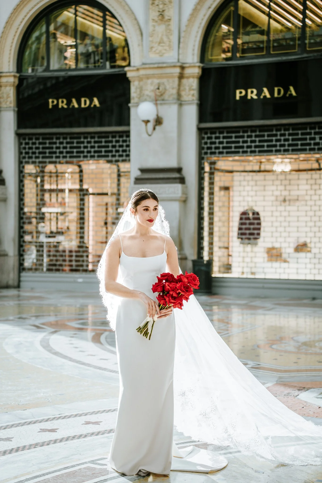 A bride in a white wedding dress holding a bouquet of red roses in a building with ornate architecture, large arched windows, and black and white tiled flooring.