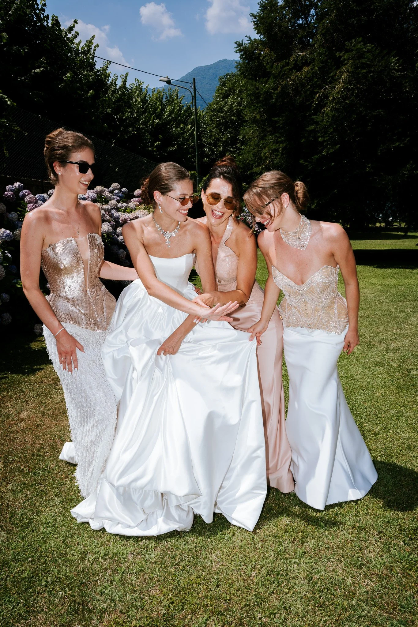 Four women in elegant dresses laughing and smiling outdoors on a grassy area with trees and mountains in the background.