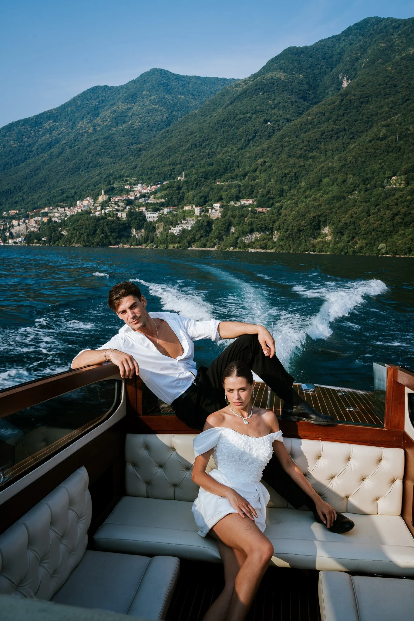 Two young adults, a man and a woman, relaxing on a boat during daytime with a large green mountain and a small hillside town in the background. The woman is wearing a white dress with off-the-shoulder sleeves and jewelry, sitting with one leg exposed