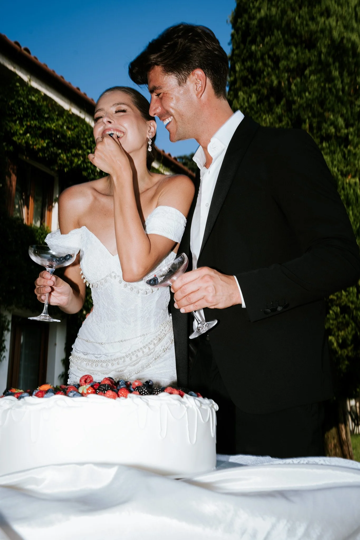 A bride and groom laughing and enjoying themselves at a wedding reception, standing next to a large white cake decorated with berries.