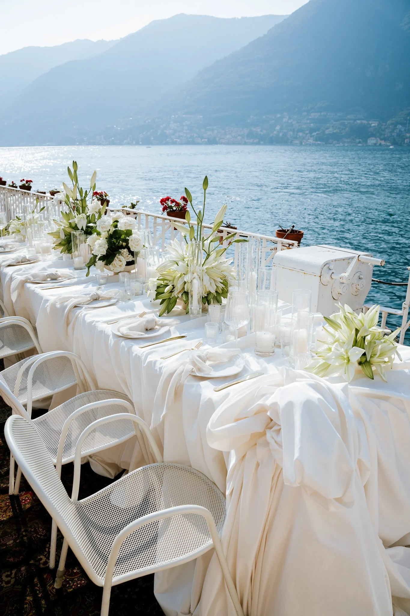 A long dining table decorated with white tablecloth, white floral arrangements, and candles, positioned outdoors overlooking a body of water and mountains in the background.