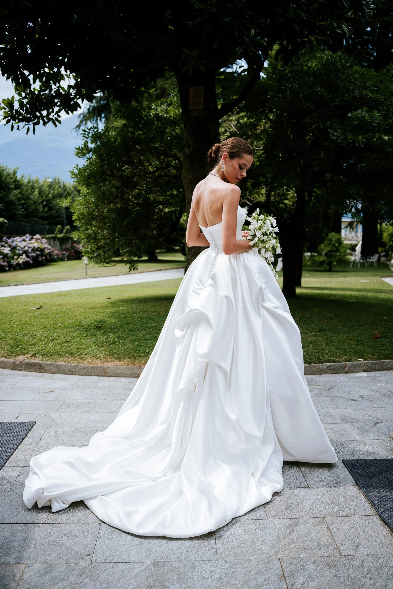A bride in a strapless white wedding gown with a long train, holding a bouquet of white flowers, standing outdoors on a paved area with green grass and trees in the background.