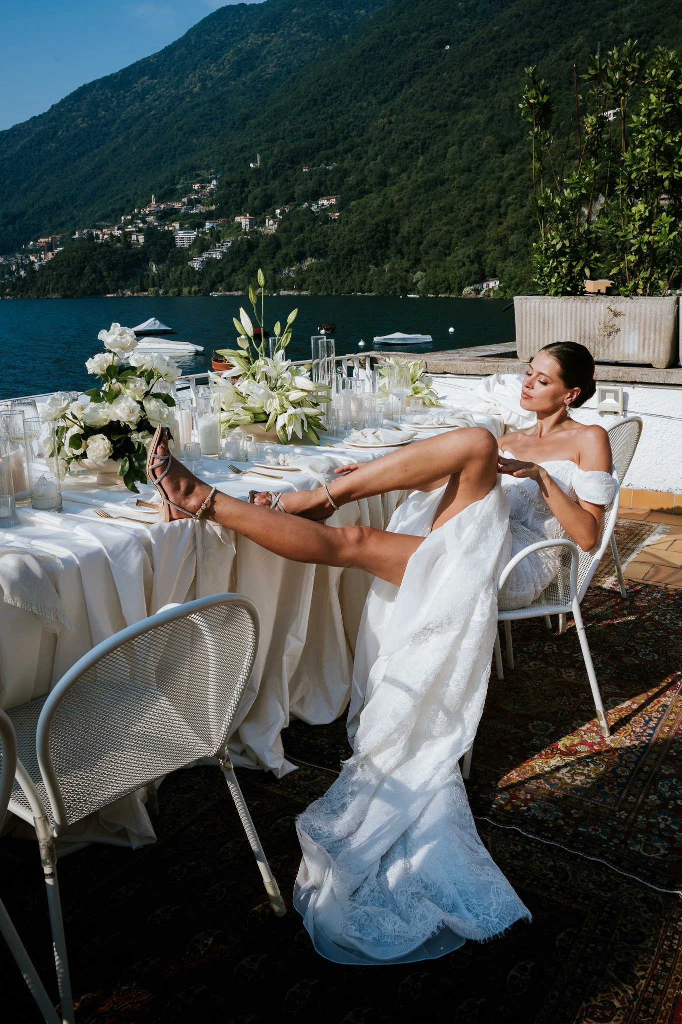 A woman in a white wedding dress relaxing on a patio with a lake and green mountain in the background, sitting on a white chair with golf shoes and a bracelet.
