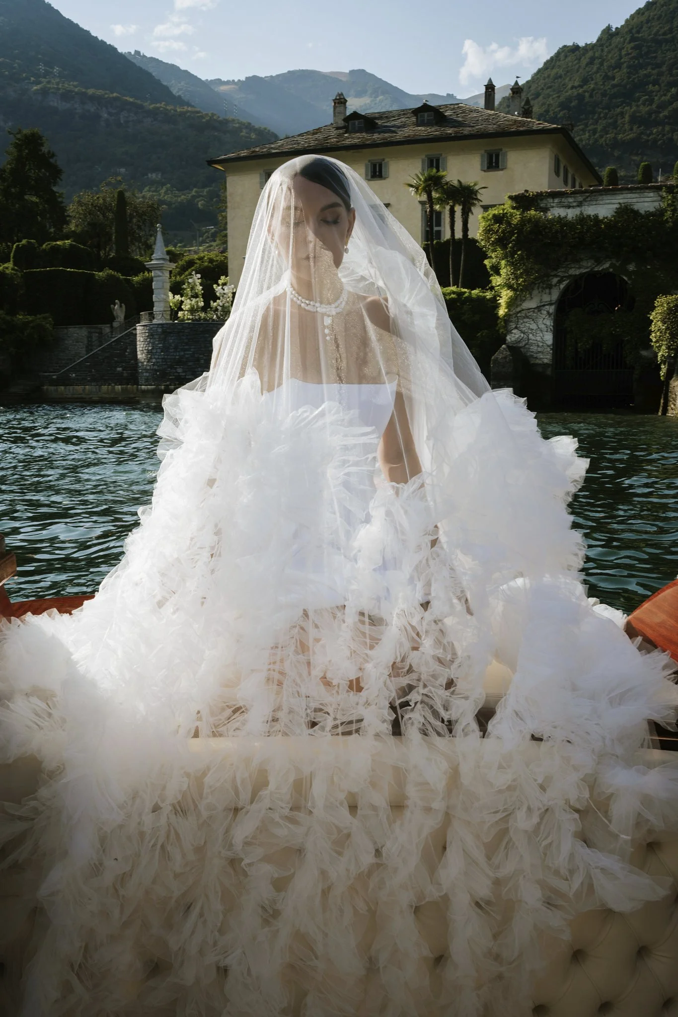 Bride in a veil and dress with a long train standing by the water at Villa del Balbiano, Lake Como, with the villa and mountains behind her.