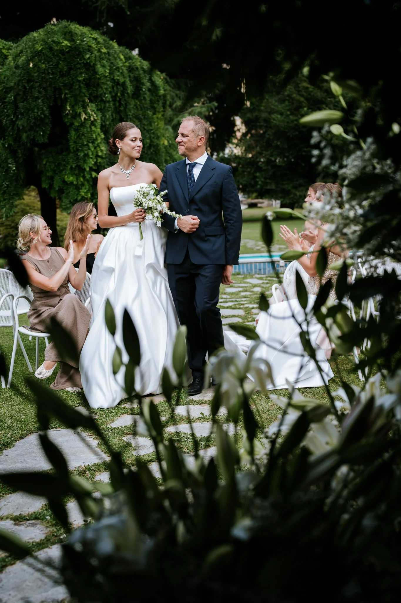 A bride in a white wedding gown holding a bouquet, walking with a groom in a dark suit during an outdoor wedding ceremony, surrounded by seated guests and lush greenery.
