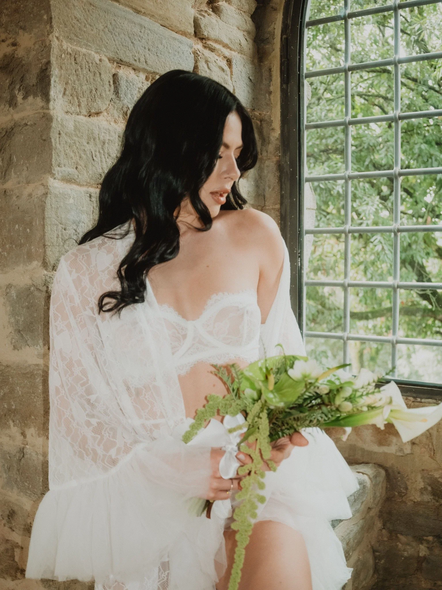 A woman with long black hair in a white lace dress holding a bouquet of green and white flowers near a window with a stone wall behind her.