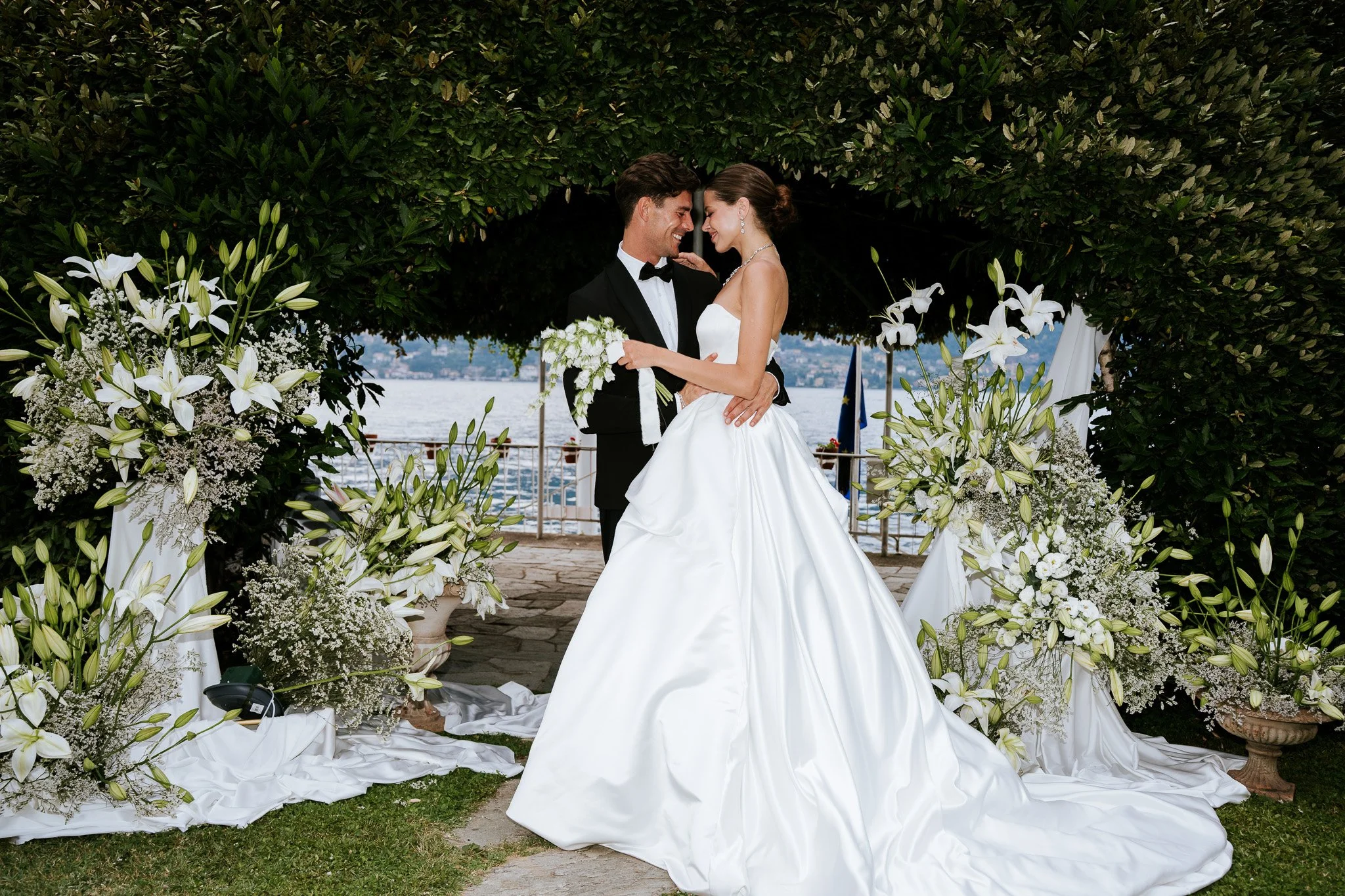 A bride and groom smiling and touching foreheads in a wedding arch decorated with white lilies and greenery, near a body of water.