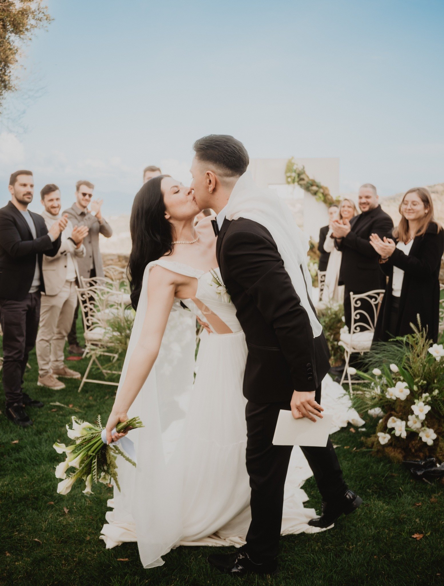 A bride and groom kiss during their outdoor wedding ceremony, with guests clapping and smiling in the background, on a grassy area under a cloudy sky.
