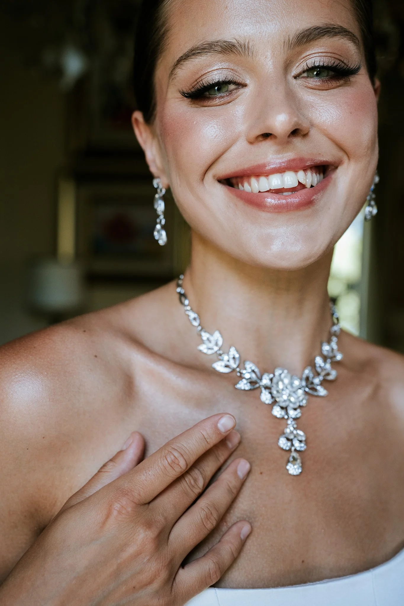 Close-up of a woman wearing a silver floral diamond necklace and matching earrings, smiling, with her left hand resting on her chest, and her bare shoulder visible.