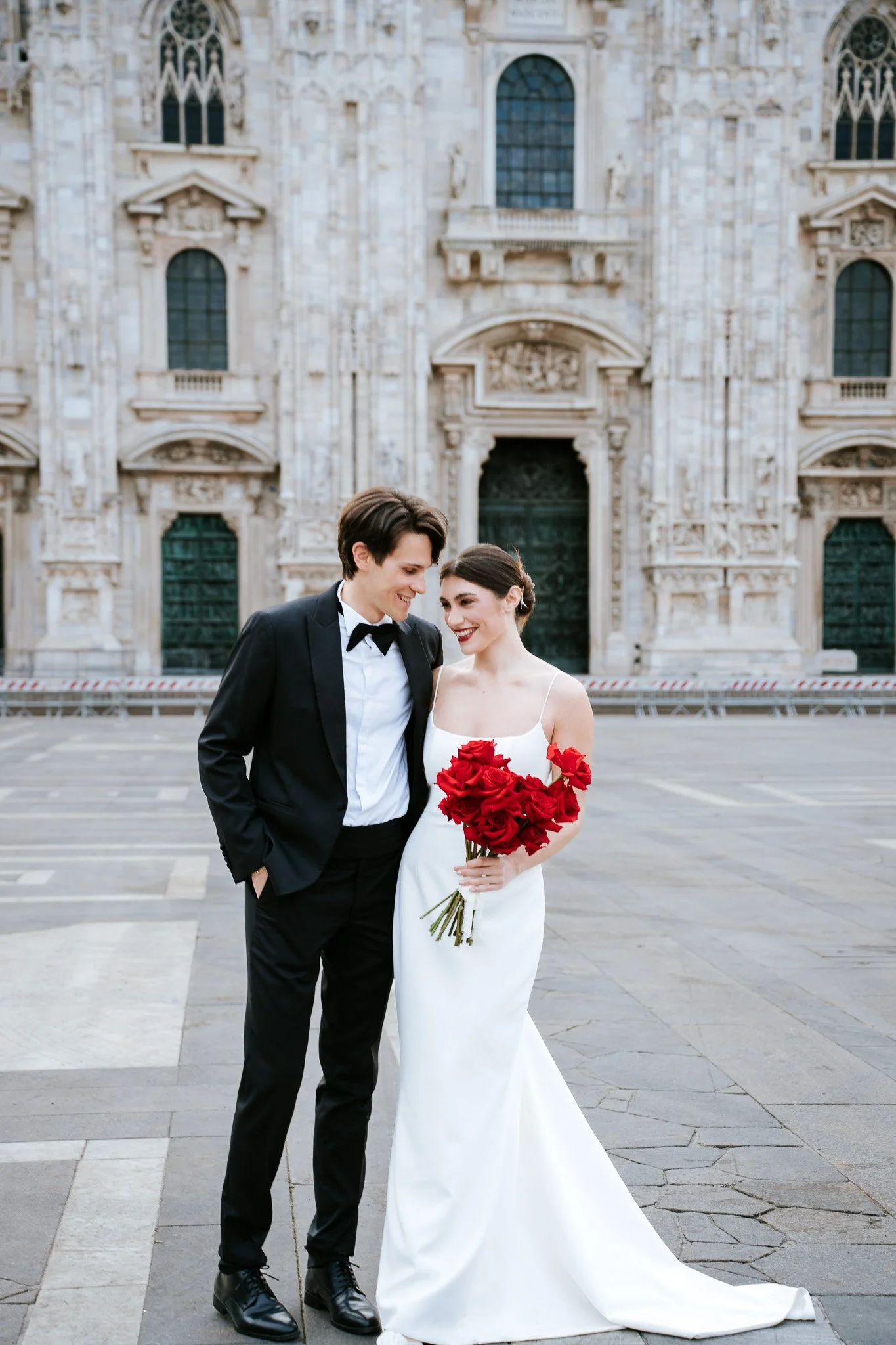 A newlywed couple stands close together in front of a historic building, with the groom dressed in a black tuxedo with a bow tie and the bride wearing a white wedding gown and holding a bouquet of red roses. They are smiling and looking at each other