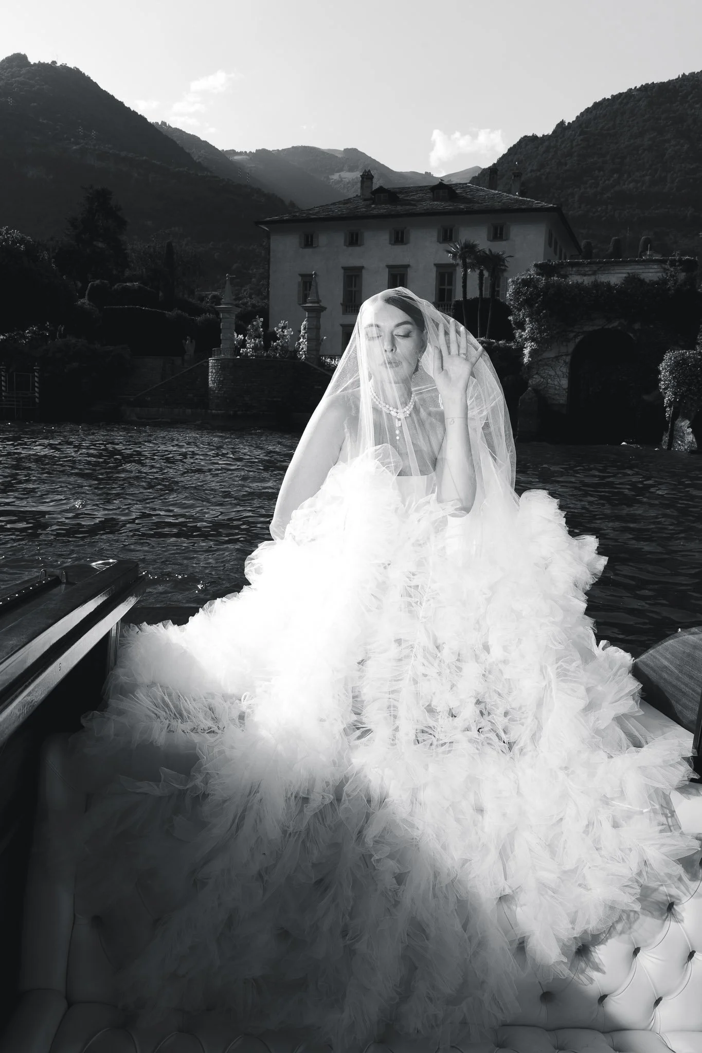 Black and white photo of a bride in her veil on a boat on Lake Como, with Villa del Balbiano and the mountains behind her.