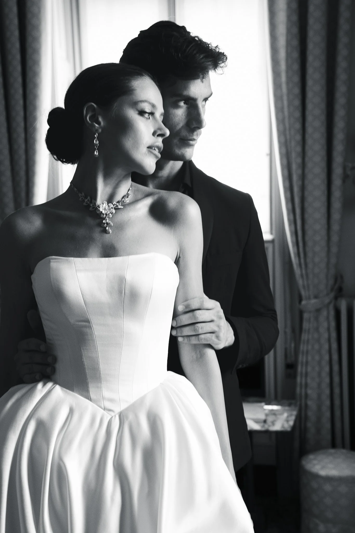A couple dressed in formal attire, with the woman wearing a white strapless gown and jewelry, and the man in a dark suit, standing closely together near a window with curtains.