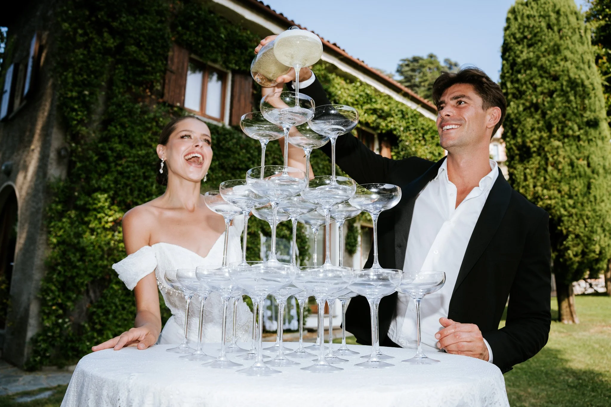A newlywed couple building a tower of empty champagne glasses at an outdoor wedding reception, smiling and celebrating.