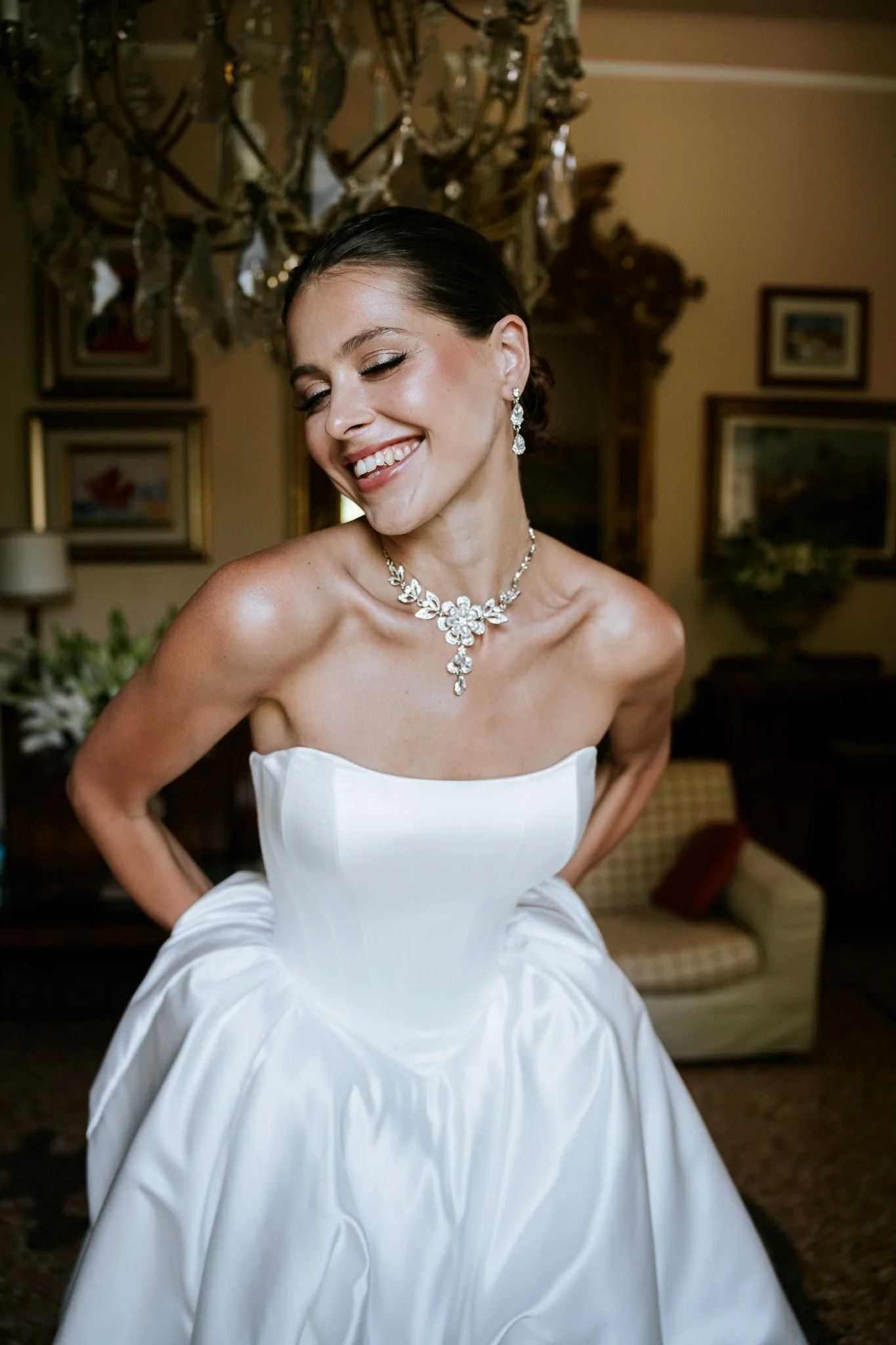 A woman dressed in a white strapless wedding gown, smiling with eyes closed, wearing diamond jewelry, standing in a decorated room with artworks and a chandelier.