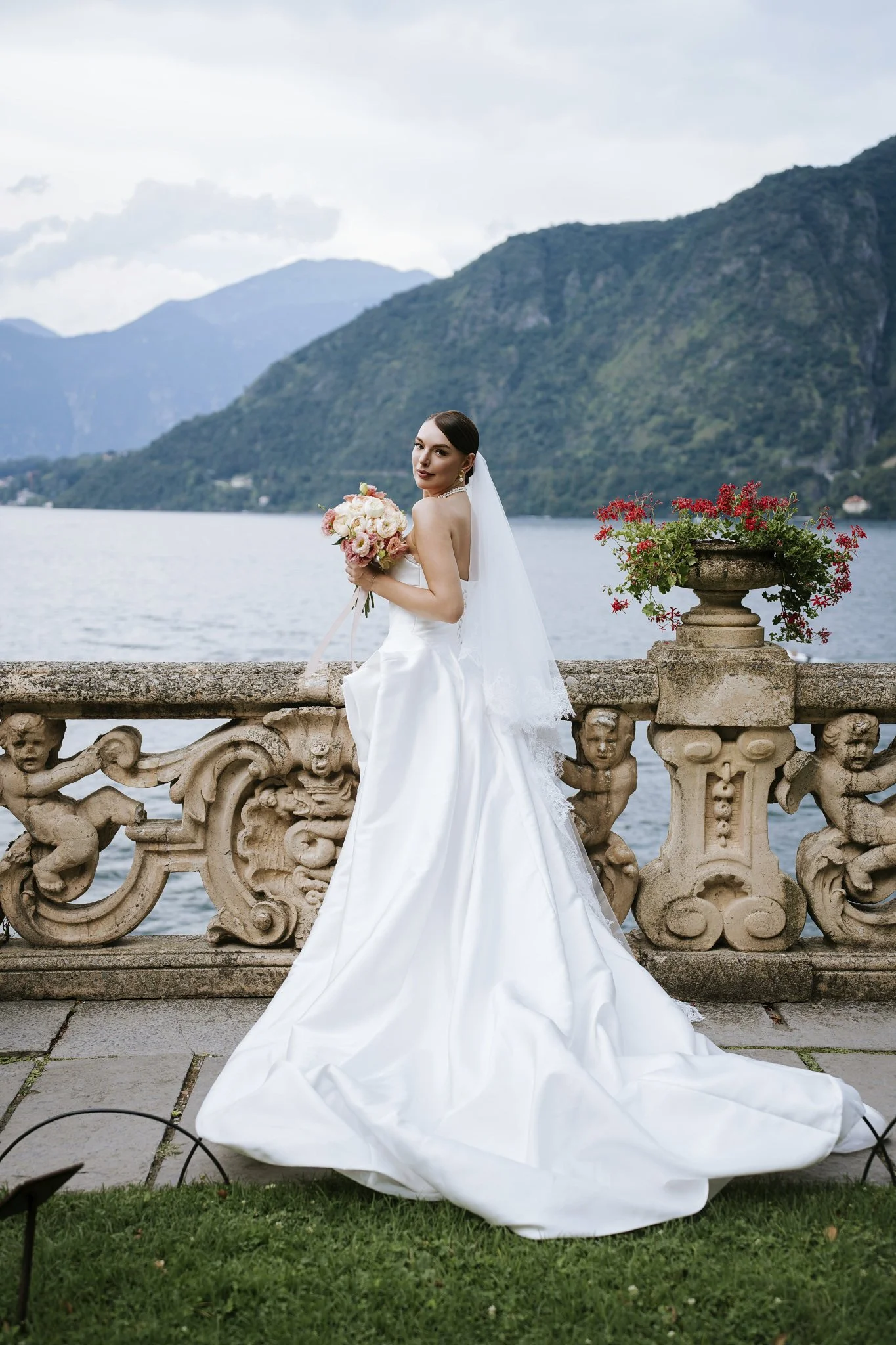 Bride holding a bouquet of pink, white, and cream flowers on the terrace at Villa del Balbianello, Lake Como, with lake and mountain views behind her.