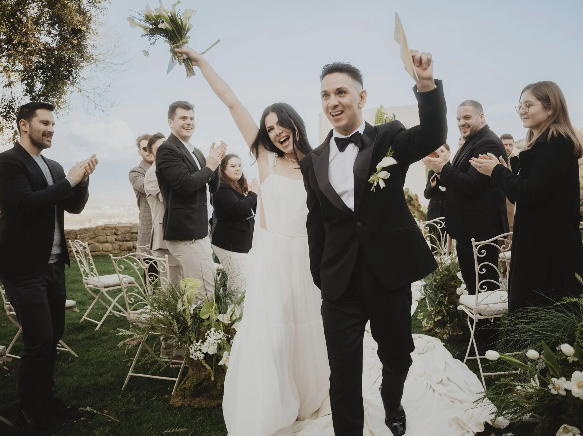 Bride raising her bouquet as the groom and guests celebrate after an outdoor wedding ceremony.