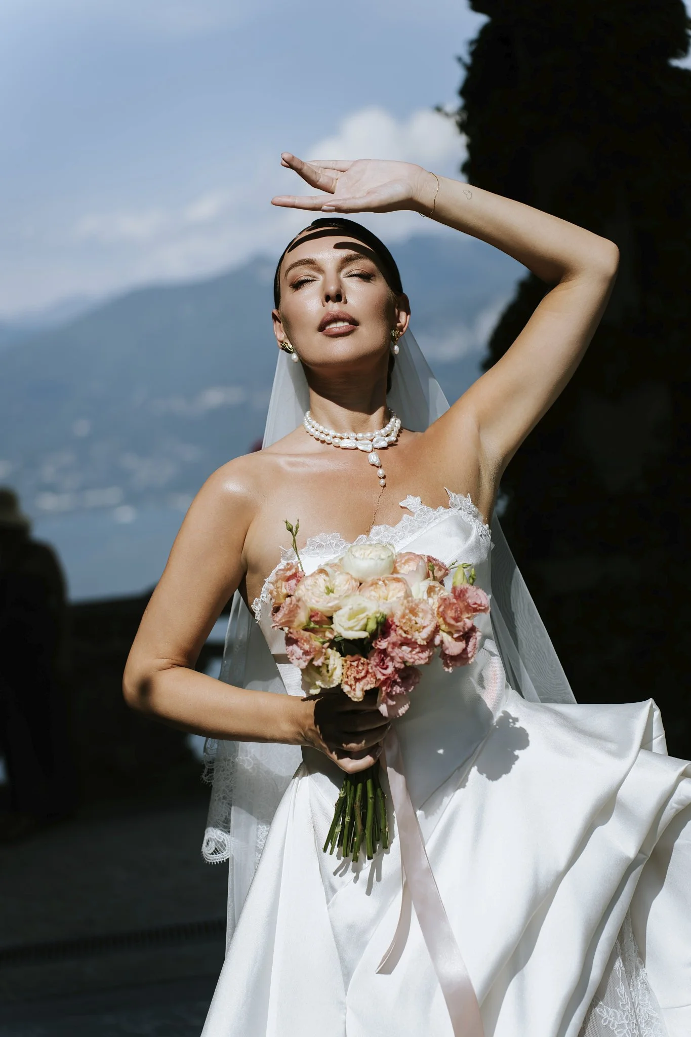 Bride shielding her eyes from the sun at Villa del Balbianello, Lake Como, against a mountain backdrop.