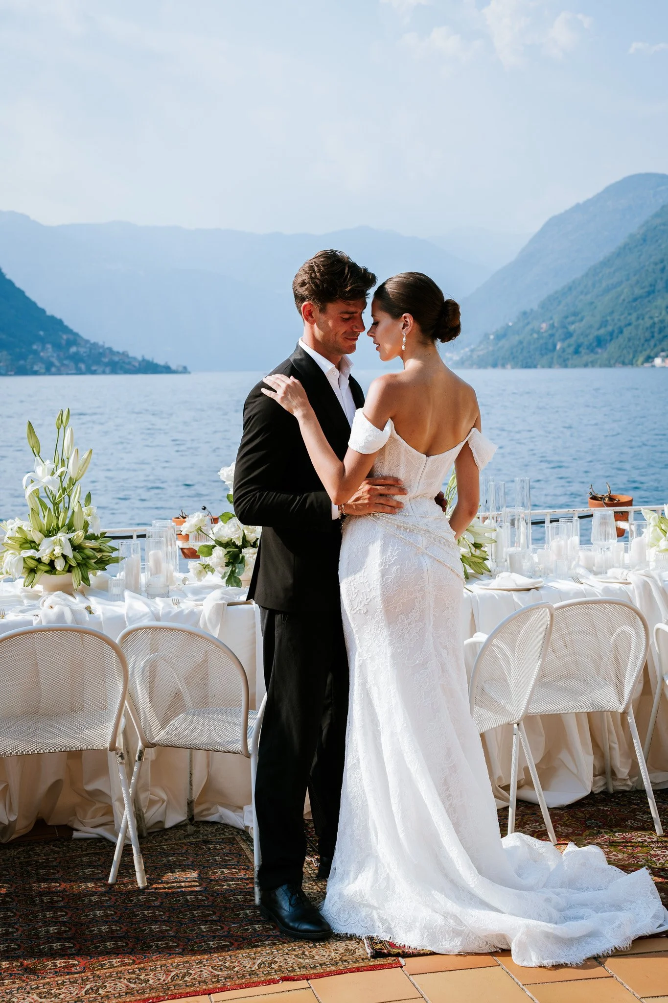 A bride and groom standing close together, embracing during their wedding ceremony by a lakeside with mountains in the background, decorated with white flowers and candles on a long table.