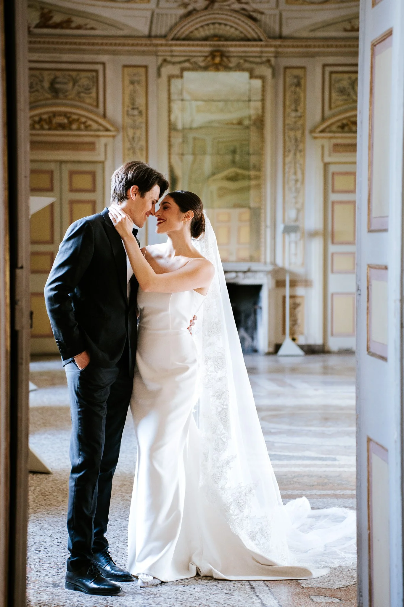 A bride and groom are standing close together in a luxurious, ornately decorated room with detailed wall and ceiling art. They are touching foreheads and smiling warmly at each other; the bride has her hand on the groom's face. The groom is dressed i