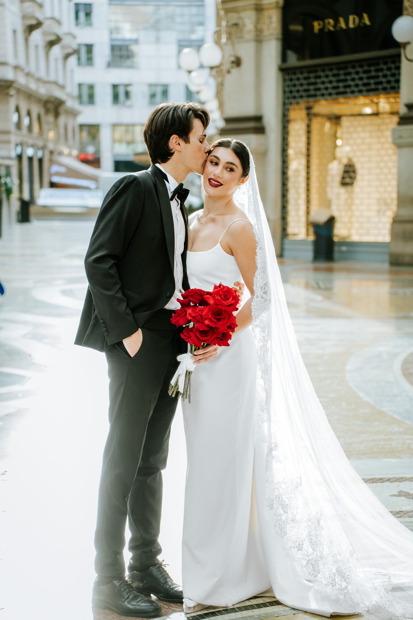 A smiling bride with dark hair in an elegant white wedding dress holding a bouquet of red roses, and a groom in a black tuxedo kisses her on the cheek outdoors in an urban setting.
