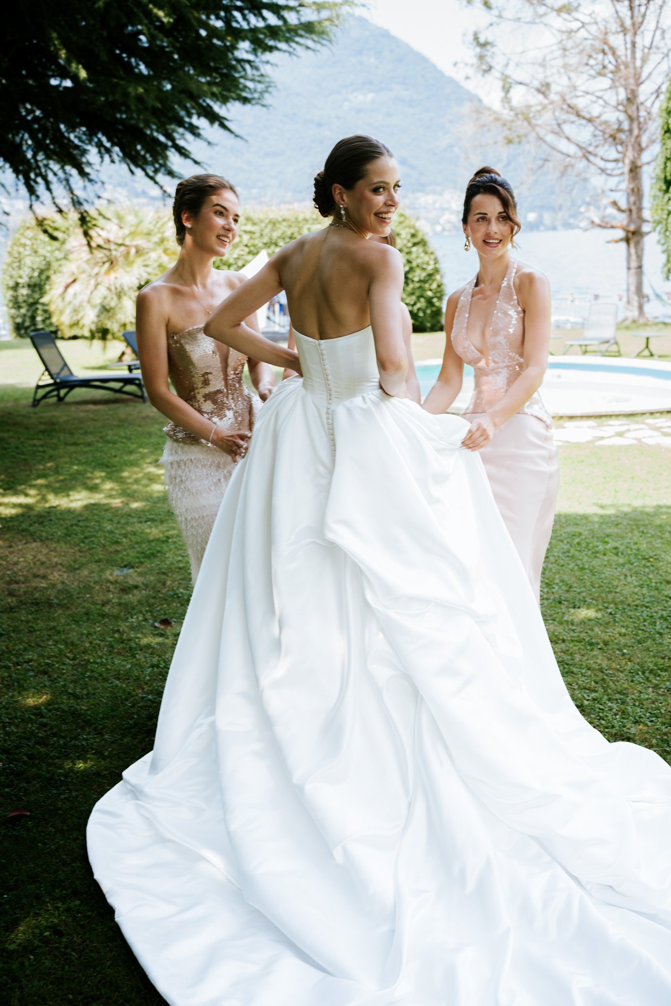 Four women dressed in formal attire standing outdoors near a body of water with mountains in the background.
