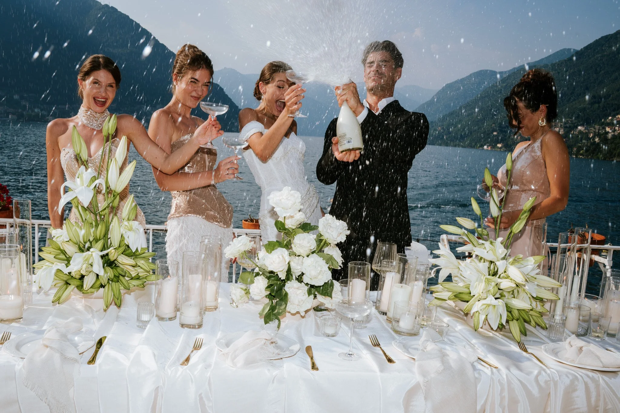 A group of five people celebrating at a wedding reception outdoors by the water, with mountains in the background, as they enjoy champagne and laughter.