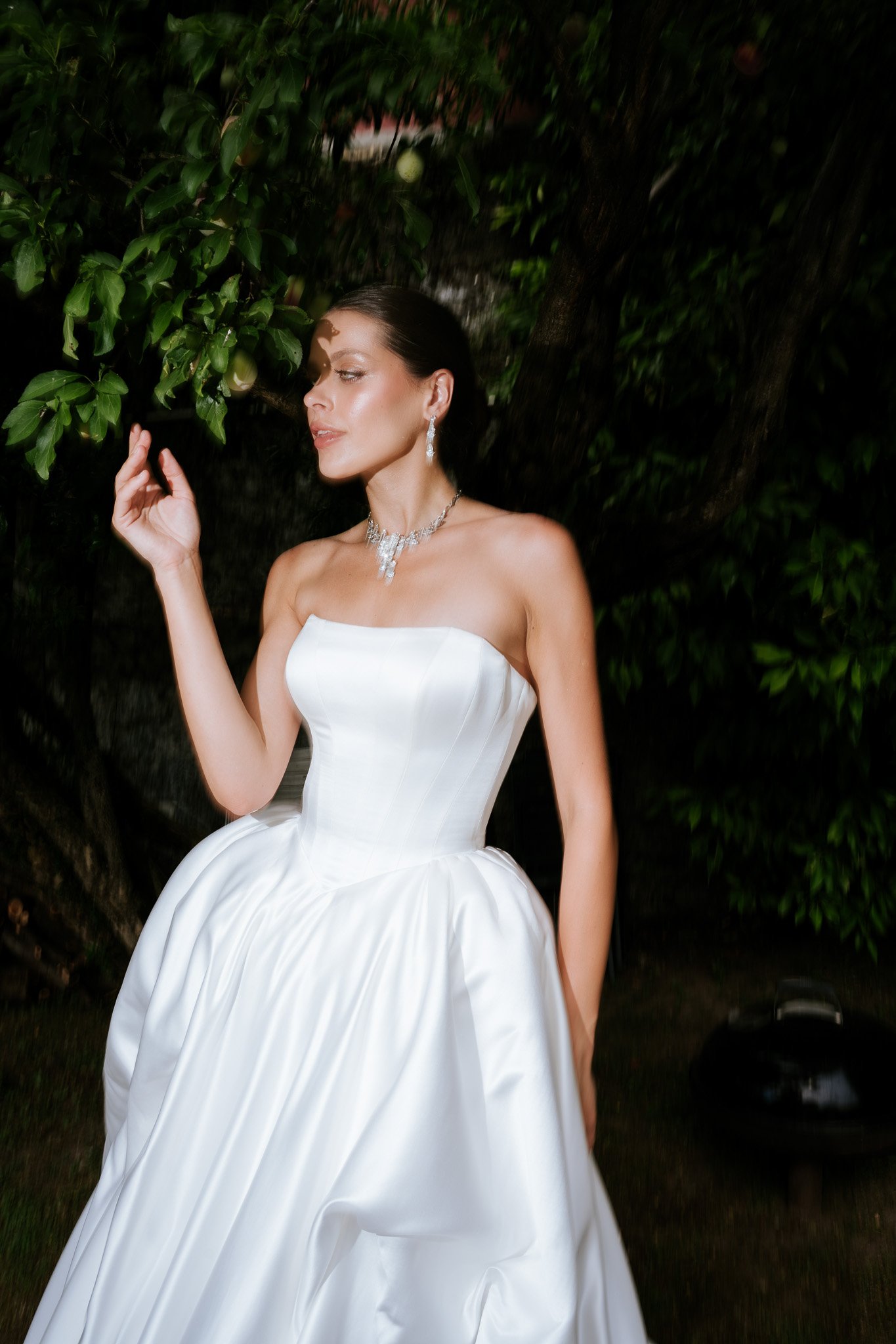 A woman wearing a white strapless wedding dress and silver jewelry standing outdoors near dark green leafy trees.