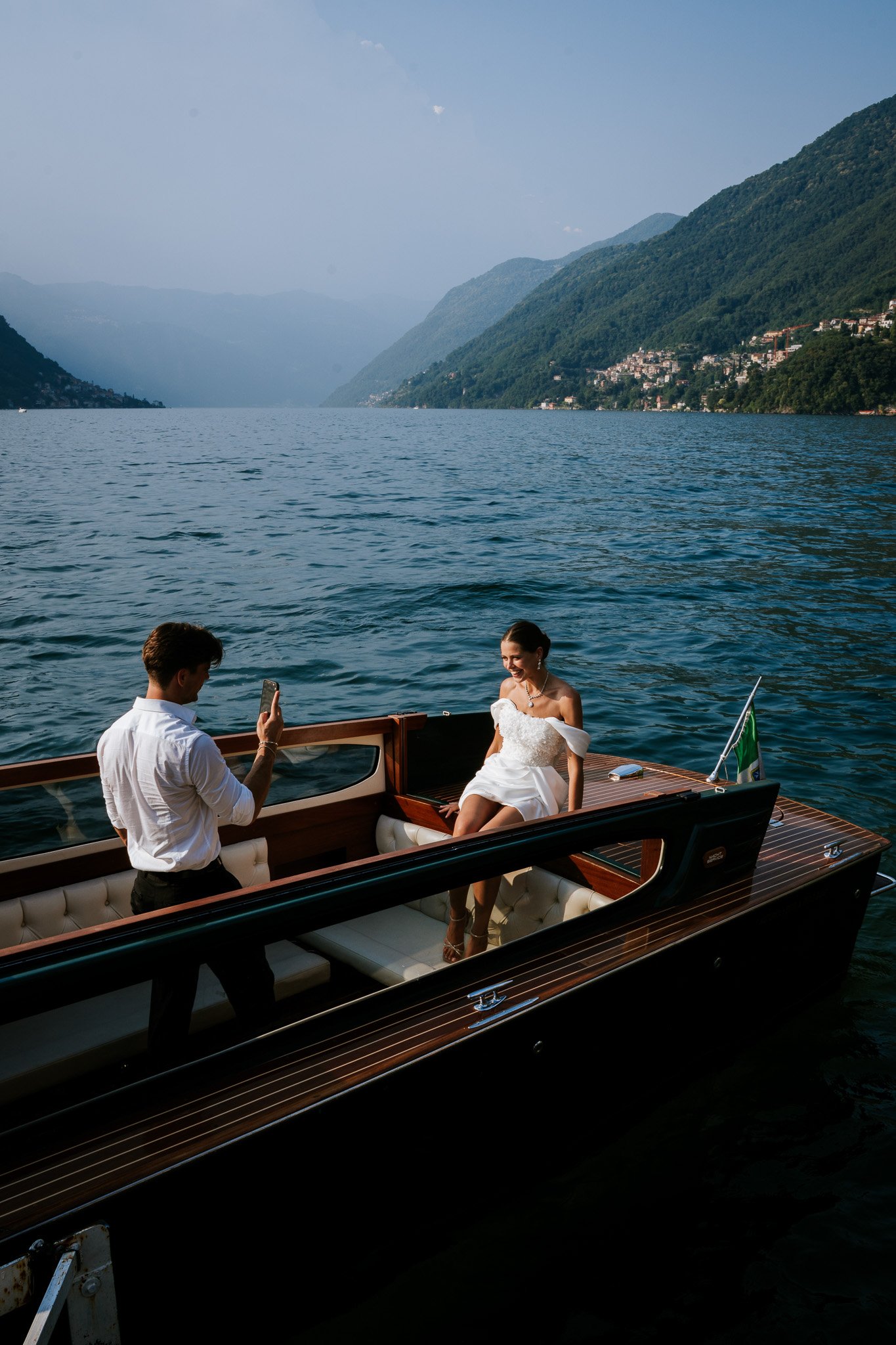 A woman in a white dress sitting on a boat while a man in a white shirt takes her photo during the daytime, with a large lake, green hills, and mountains in the background.