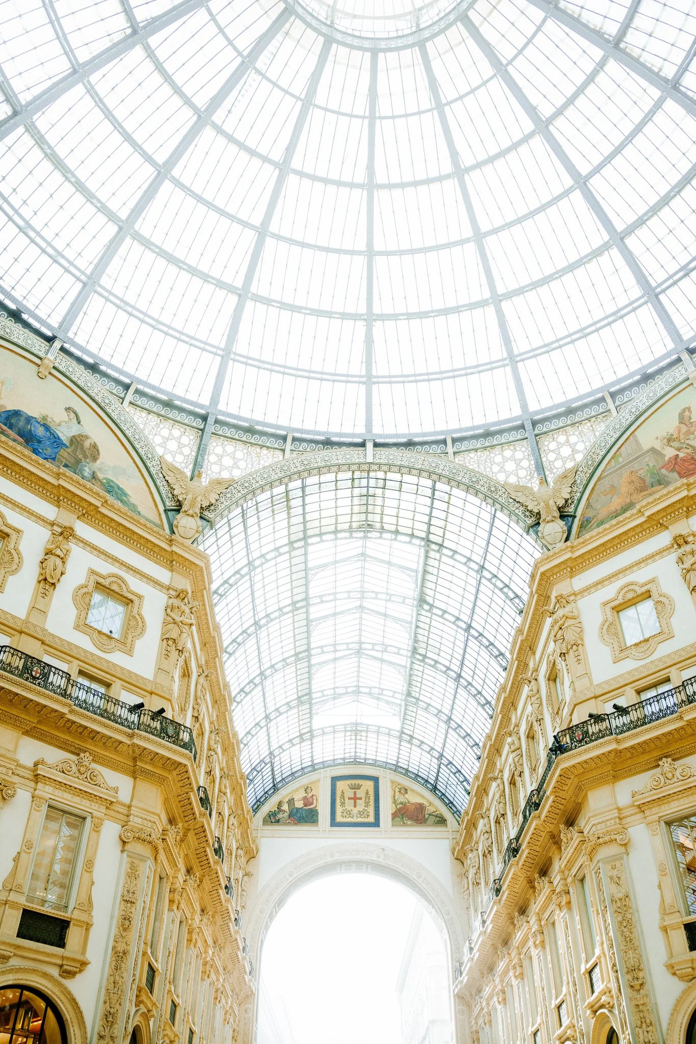 Interior of a grand shopping arcade with a large glass-domed ceiling, ornate cream-colored walls, and decorative architectural details.