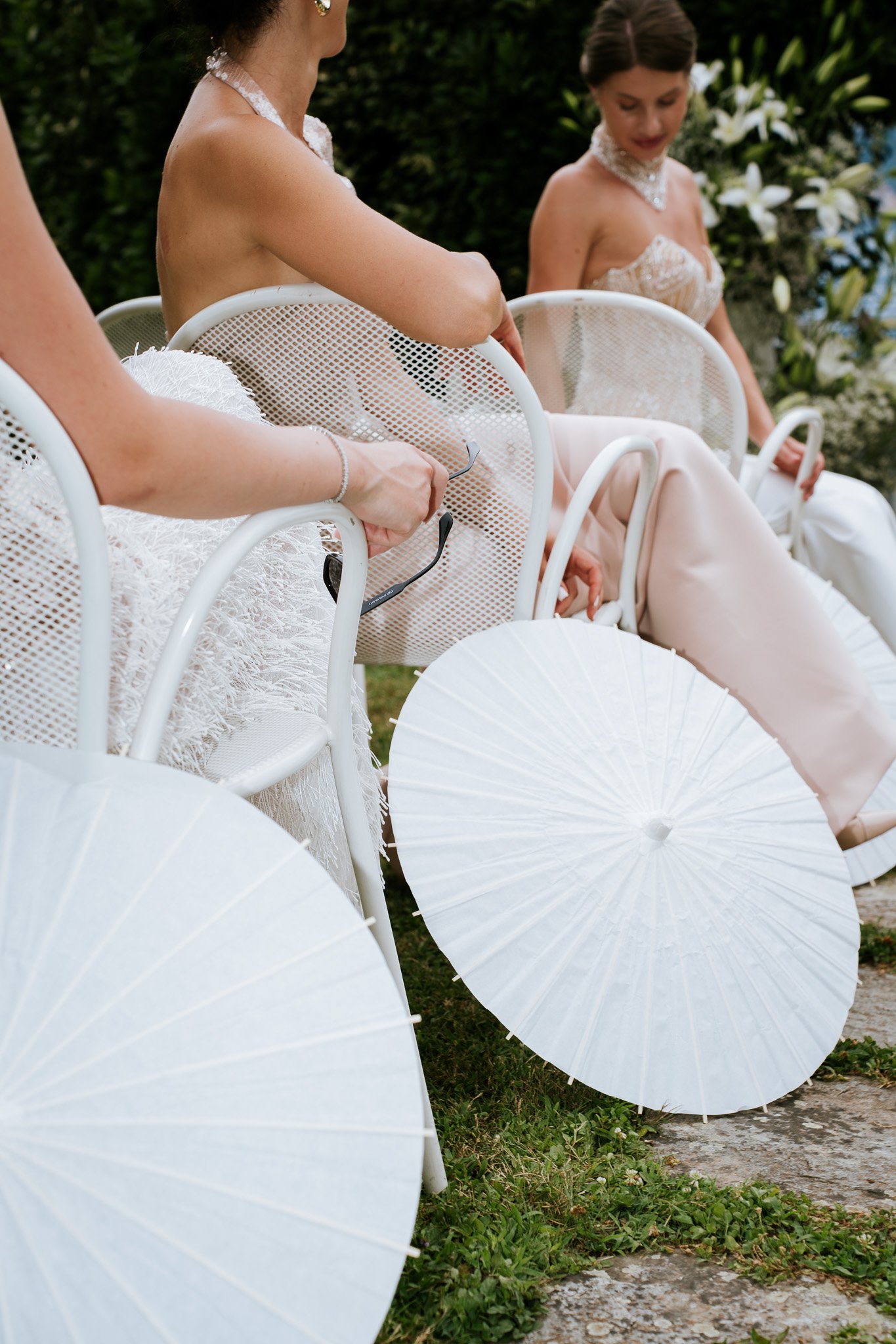 Several white parasols arranged on a grassy surface and a stone-paved path, with three women sitting on white chairs, wearing elegant dresses and holding white parasols, surrounded by greenery and white flowers.
