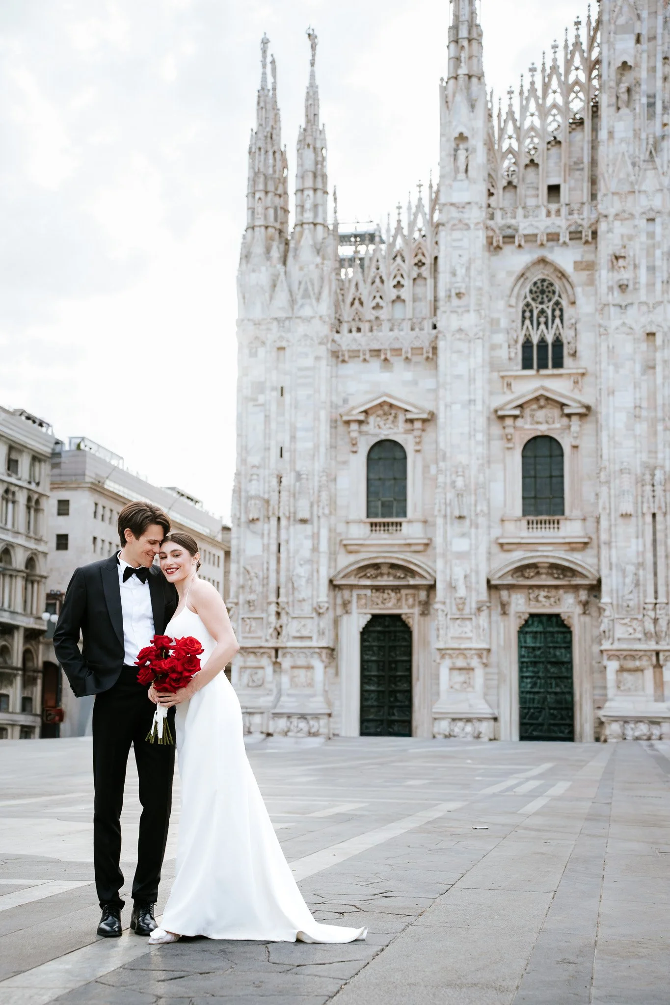 A couple in wedding attire standing close together outdoors in front of a historical building, with the woman holding a bouquet of red roses.