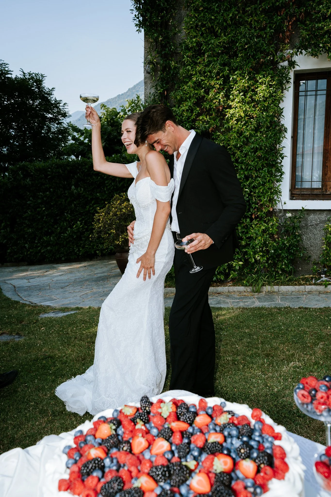 A bride and groom celebrating at a wedding reception outdoors, with a large berry and fruit dessert in the foreground. The bride is raising a glass and laughing, and the groom is leaning towards her, also smiling.