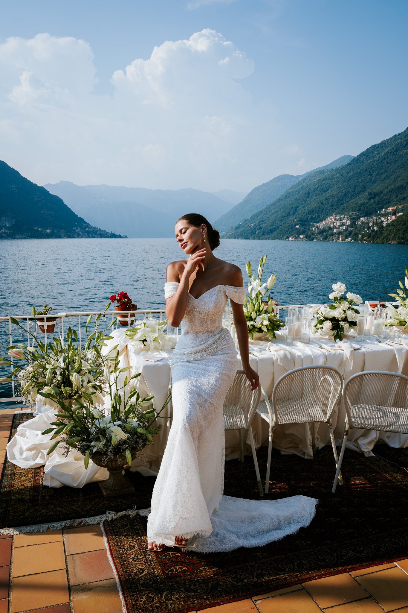 A woman in a white lace wedding dress stands on a decorative rug in front of a long table decorated with white flowers, candles, and greenery, with a mountain lake view in the background.