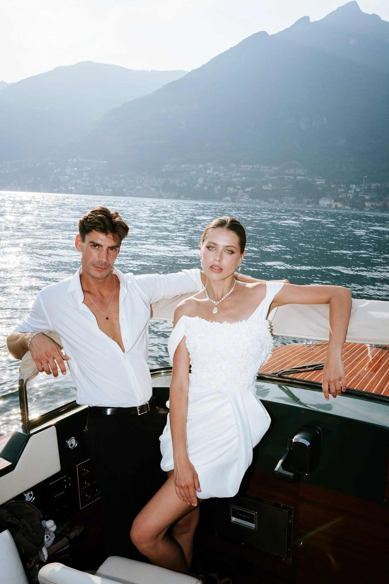 A young man and woman on a yacht, with a lake and mountains in the background. The man is dressed in a white shirt and black pants, and the woman is wearing a white dress and jewelry. The woman leans back against the yacht's railing, and the man has 