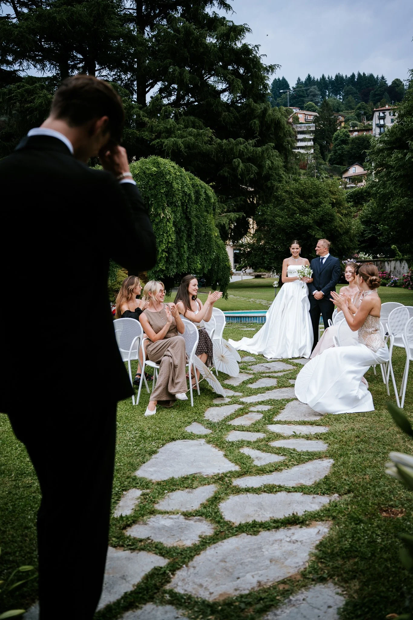 A wedding ceremony outdoors with a bride in a white gown and a groom in a navy suit, standing in front of seated guests clapping, with officiant in the background and green trees and houses on a hillside.