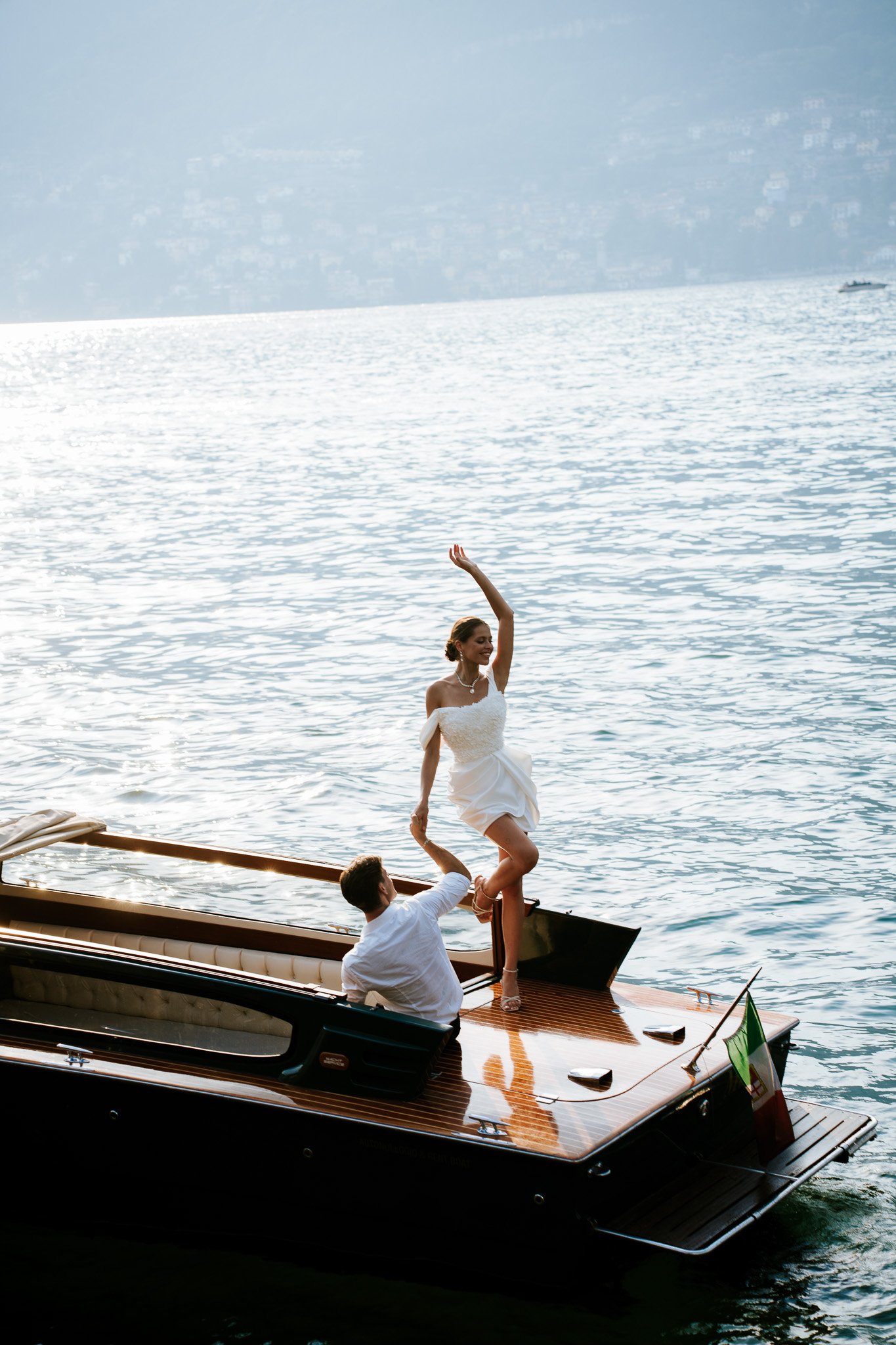 A woman in a white wedding dress standing on a boat, holding hands with a man in a white shirt, with the ocean and a distant shoreline in the background.