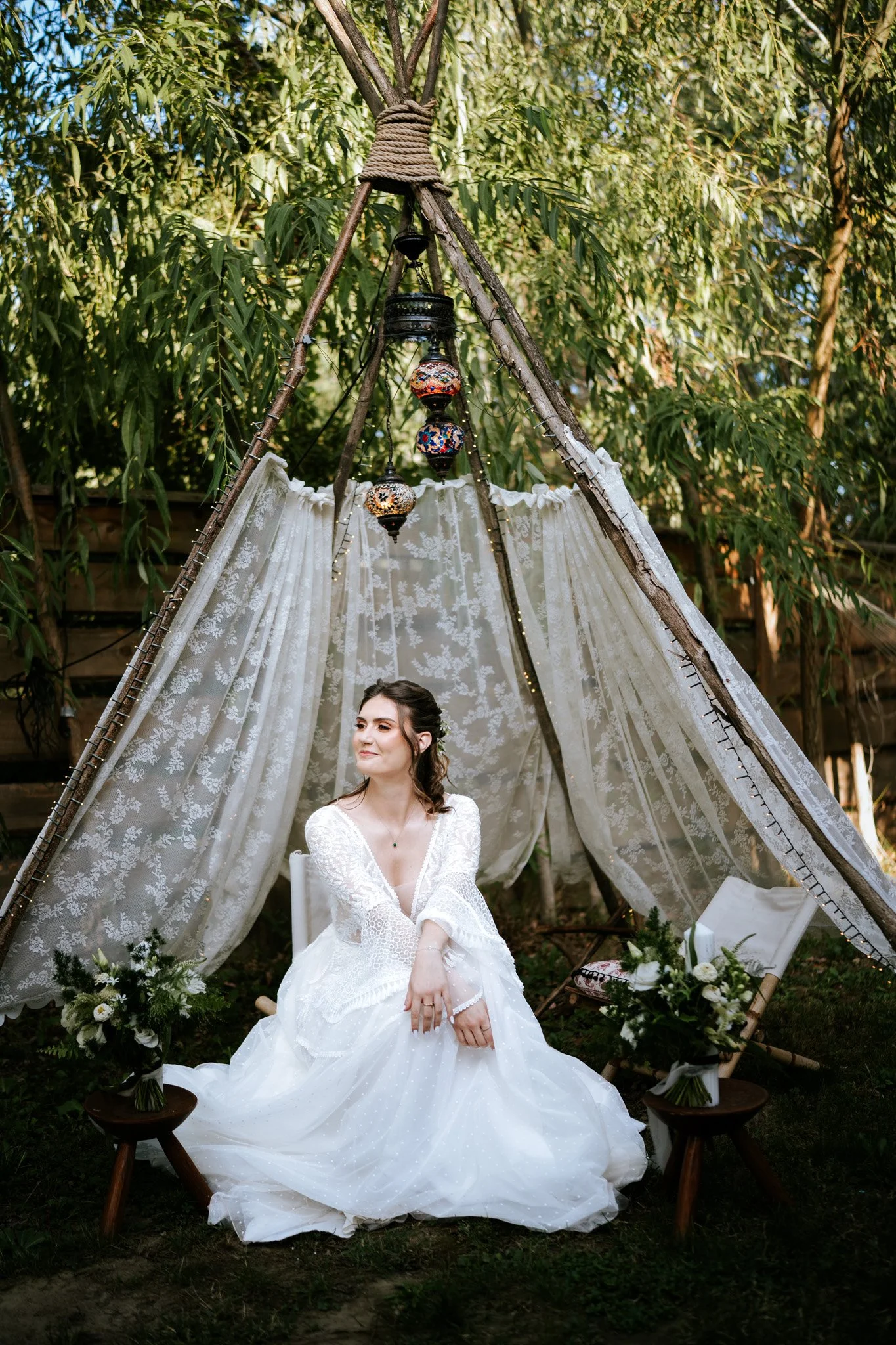 A woman wearing a white bridal dress seated outdoors under a canopy made of rustic sticks and lace fabric, with hanging colorful Moroccan-style lanterns above her. The area is decorated with flowers and greenery.