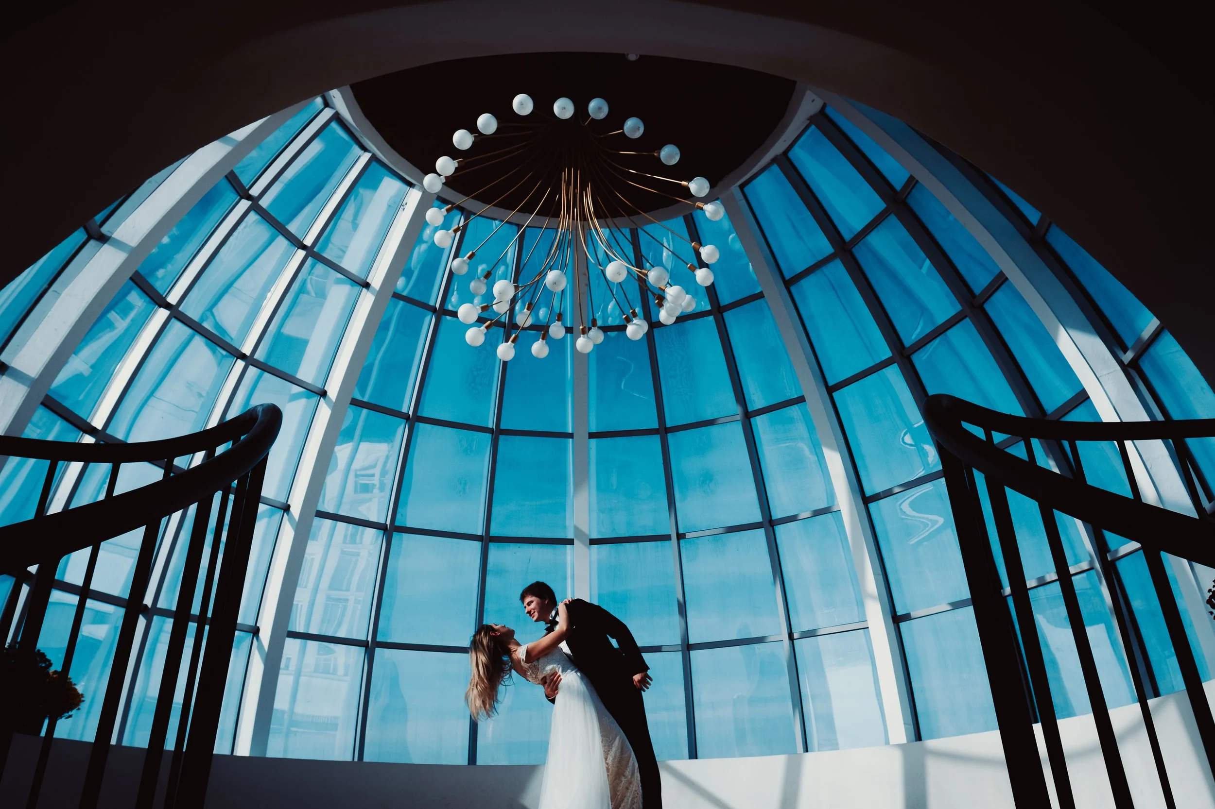 Bride and groom dancing beneath a circular glass ceiling and chandelier during a luxury wedding celebration.