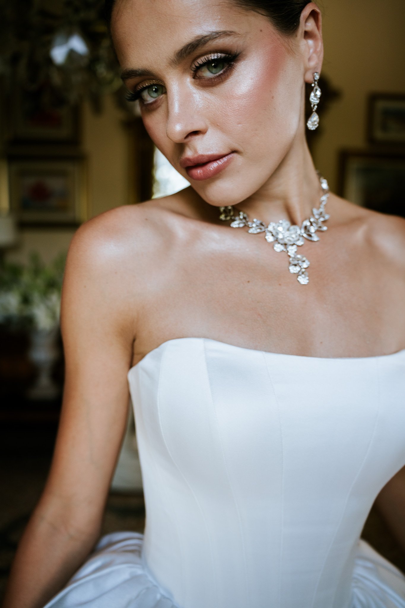 A woman in a white strapless dress wearing diamond jewelry, including earrings and a necklace, with a blurred background of a room with framed pictures and plants.