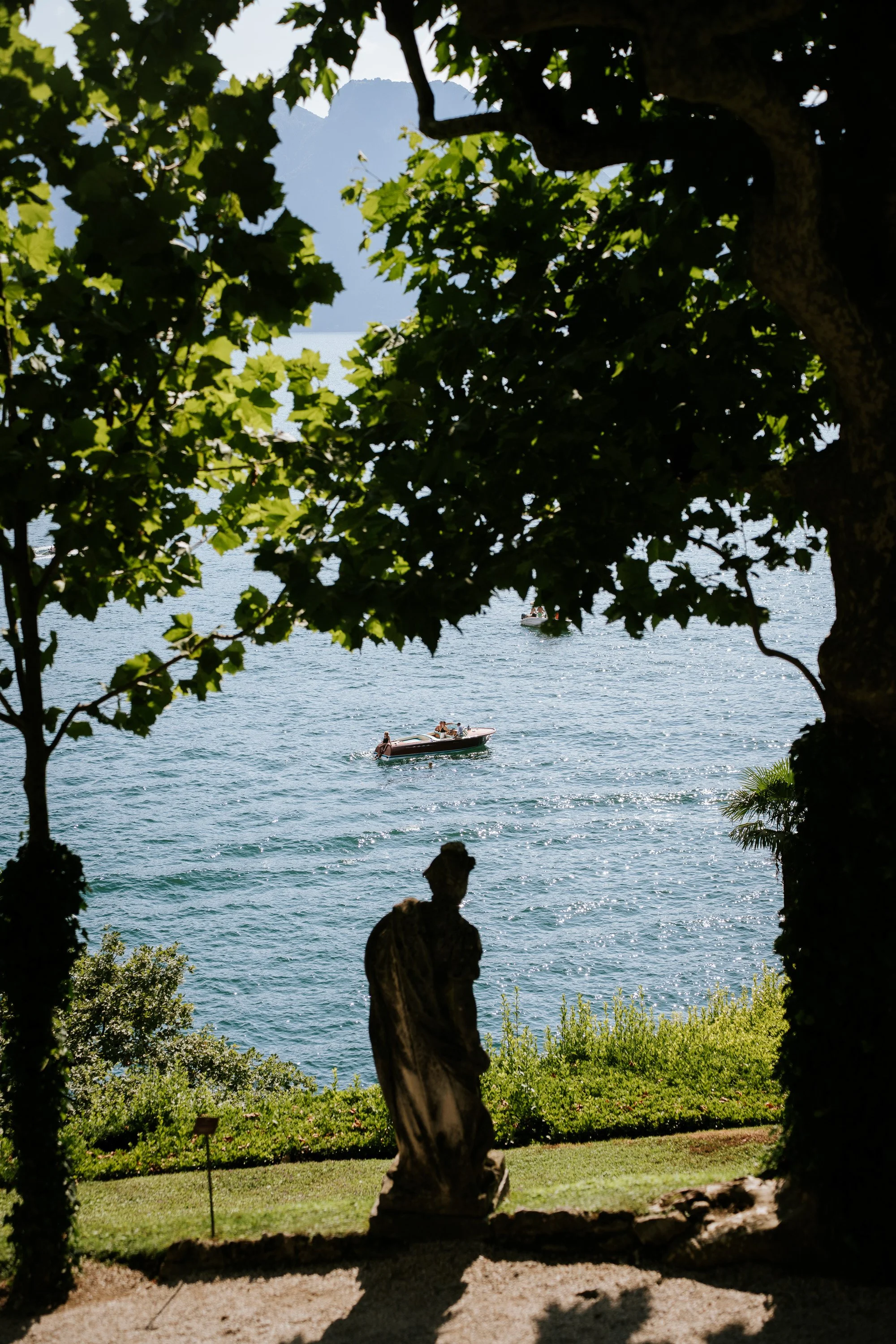 View of Lake Como from the gardens of Villa del Balbianello, framed by trees with a statue in the foreground.