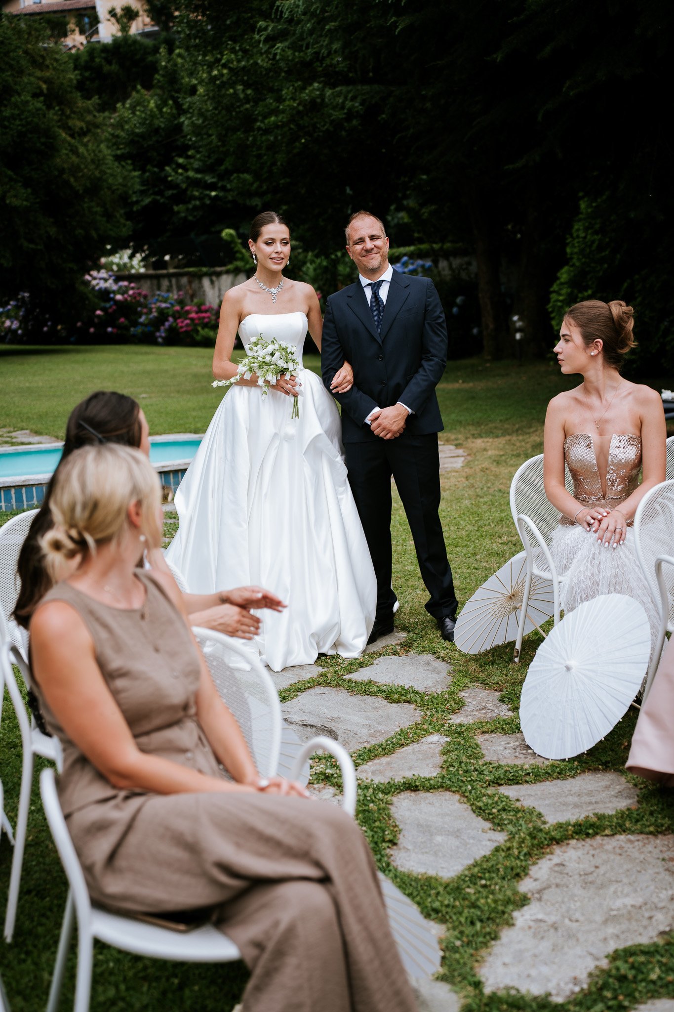 A bride and her father standing outdoors during a wedding ceremony, with seated guests around them. The bride wears a strapless white gown and holds a bouquet, and the father wears a dark suit and tie. There are white parasols on the grass and trees 